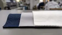 Macro photograph of three polo fabric swatches overlapping: navy honeycomb dry-fit mesh, grey polyester piqué and heavier white cotton piqué, showing the difference in knit density