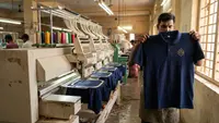 A multi-head industrial embroidery machine running navy polo shirts in a Tiruppur garment unit while an inspector holds a finished polo up to the light to check the stitching